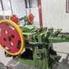 Green industrial machine with large red and yellow flywheels in a workshop, connected by white wires and metal components.
